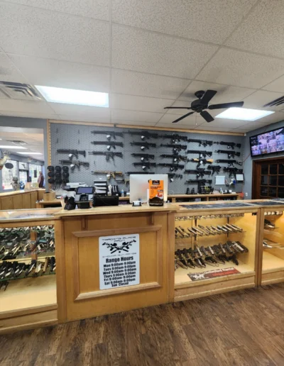A store counter with glass display cases showing handguns and a wall-mounted display of modern sporting rifles.