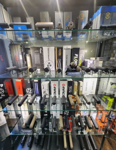 Glass display shelves showcasing a large variety of firearm suppressors from brands like Huxwrx and AAC.